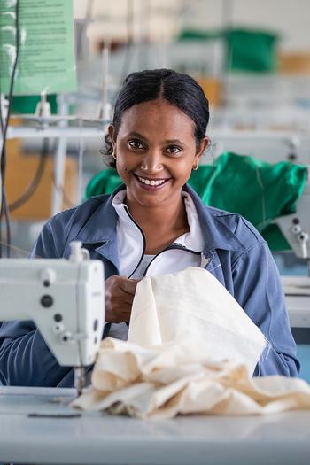 A woman sits at a sewing machine and smiles at the camera.