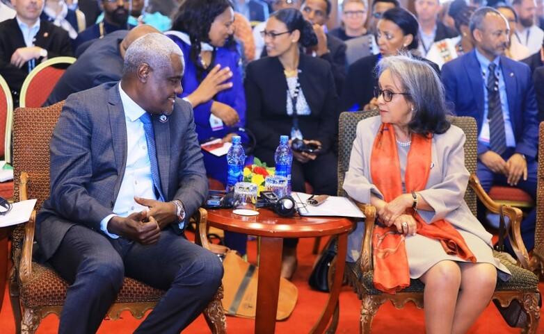 A man and woman sitting in chairs at the front of a crowded conference, engaged in discussion.
