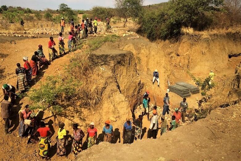 Zahlreiche Menschen einer Gemeinde bauen eine Geröllsperre, um die Fließgeschwindigkeit eines Gewässers zu verringern.
