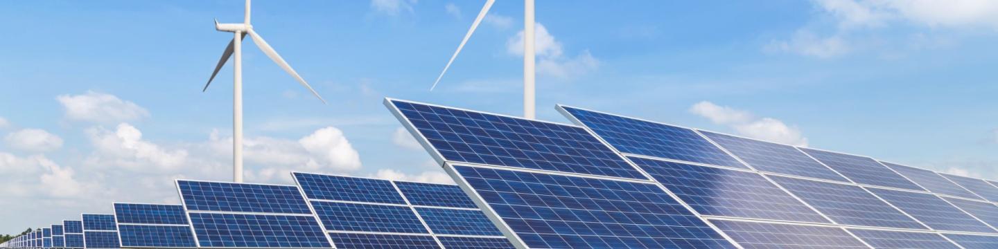 Zwei Reihen mit Solarmodulen im Vordergrund mit Windkraftanlagen vor einem hellblauen Himmel im Hintergrund stehen für die Integration der erneuerbaren Energien.