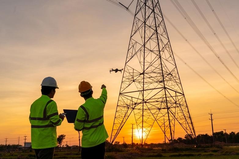 Two engineers in high-visibility jackets and hard hats use a drone to inspect a high-voltage transmission tower at sunset.