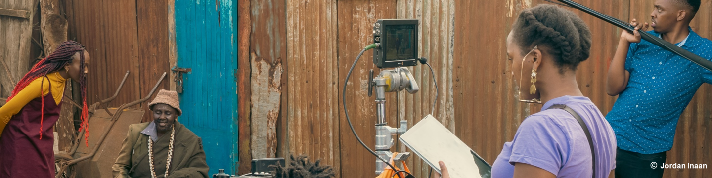 Ein Mann hält eine Tonangel und eine Frau hält ein Tablet und steht hinter einer Kamera. Vor der Kamera agieren zwei Darsteller*innen.