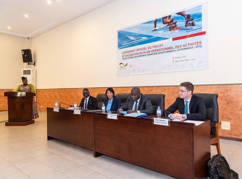 Four people seated at a long desk facing the room, in the background a woman stands at a lectern.