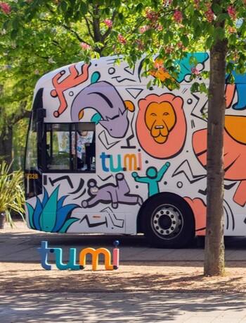 A colourful TUMI electric bus parked under trees in blossom.