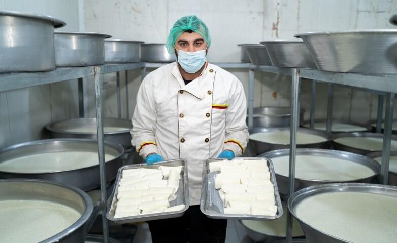 A worker in hygienic attire presents freshly made Kaymak amidst multiple large vats of heated milk in a dairy.