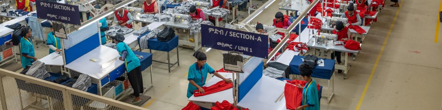 Blick von oben auf Westen tragende Arbeiter*innen, die in einer großen Bekleidungsfabrik reihenweise aufgestellte Nähmaschinen bedienen.