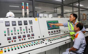 Eine Person sitzt vor einer Schalt- und Steuerungsanlage eines Industriebetriebes. Eine zweite Person in Warnweste steht neben ihr und zeigt auf einen Monitor.