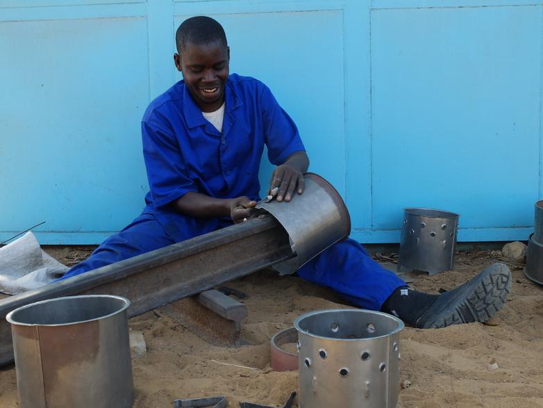 A man in blue overalls is sitting on the ground showing how ovens are made during a training session. Copyright: GIZ