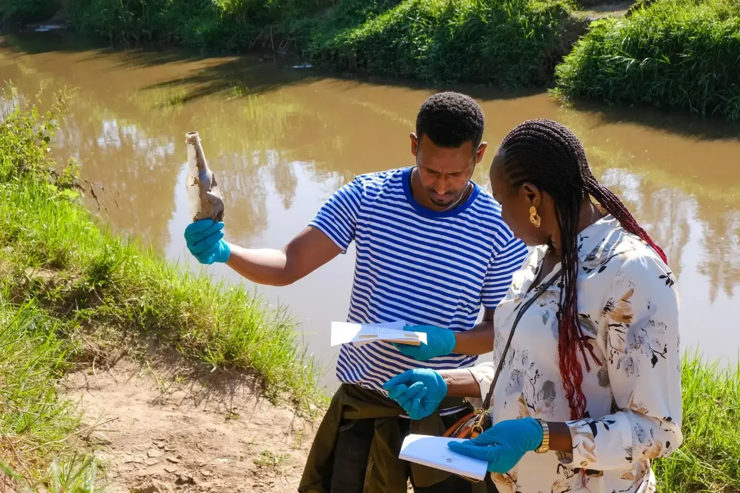 Two people with blue plastic gloves in front of a river hold paper work and a collected bottle in their hands.