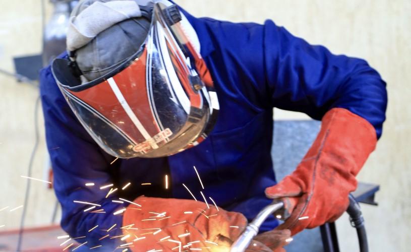 Eine Arbeitskraft in Schutzausrüstung mit Schweißhelm und Handschuhen schweißt in einer Werkstatt Metall, wobei helle Funken entstehen.