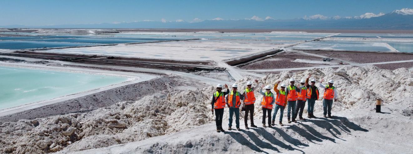 Group of people with safety helmets and bright orange safety vests standing on a hill in a salt mining area with large evaporation ponds; in the background, mountains and a clear blue sky are visible.