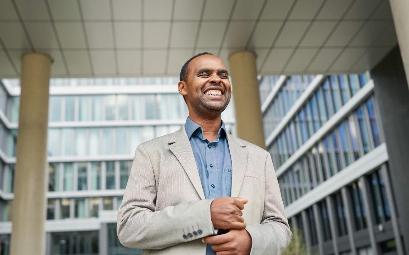 A smiling man in a light-coloured jacket and blue shirt stands in front of a modern office building with a glass façade.