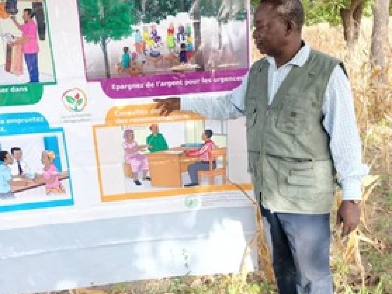 Ein Mann, der auf einem Plakat, dass unter einem Baum aufgespannt ist, etwas zeigt.