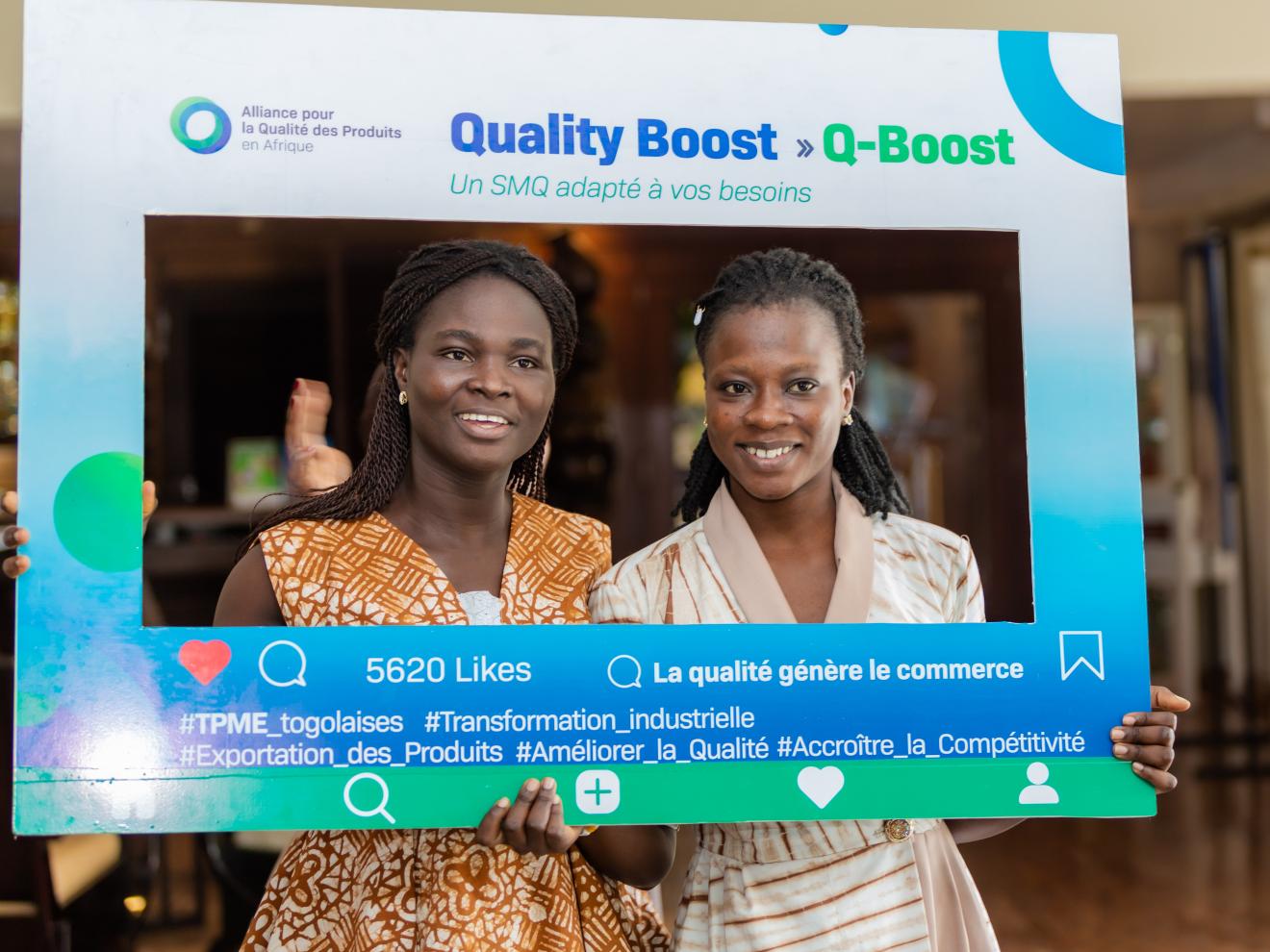 Two women smile while posing with a “Quality Boost (Q-Boost)” promotional frame.