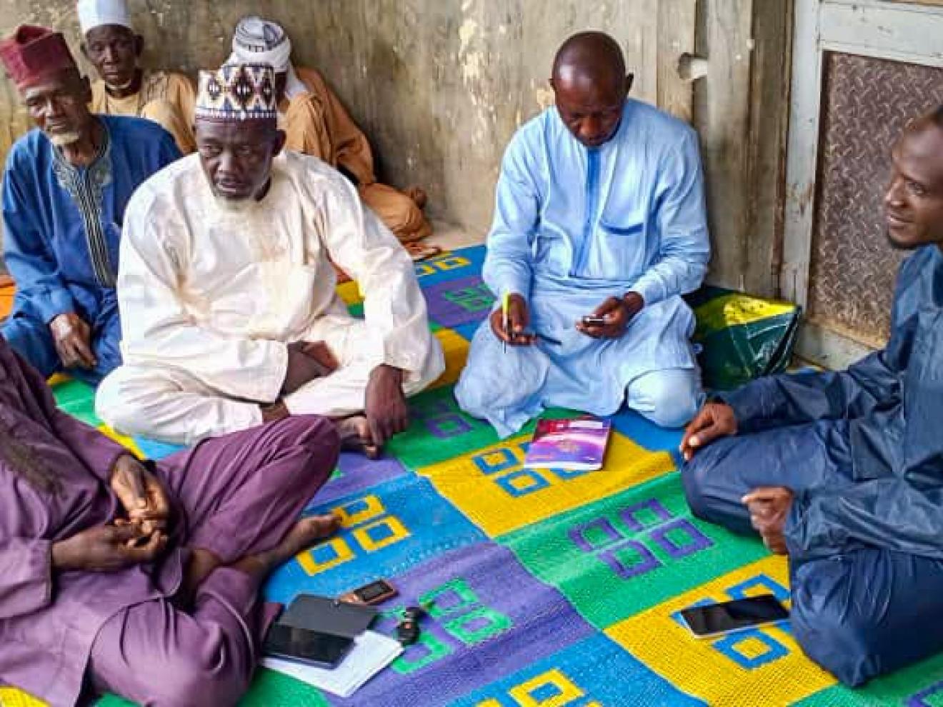 A group of men sit on a colorful mat, engaged in discussion