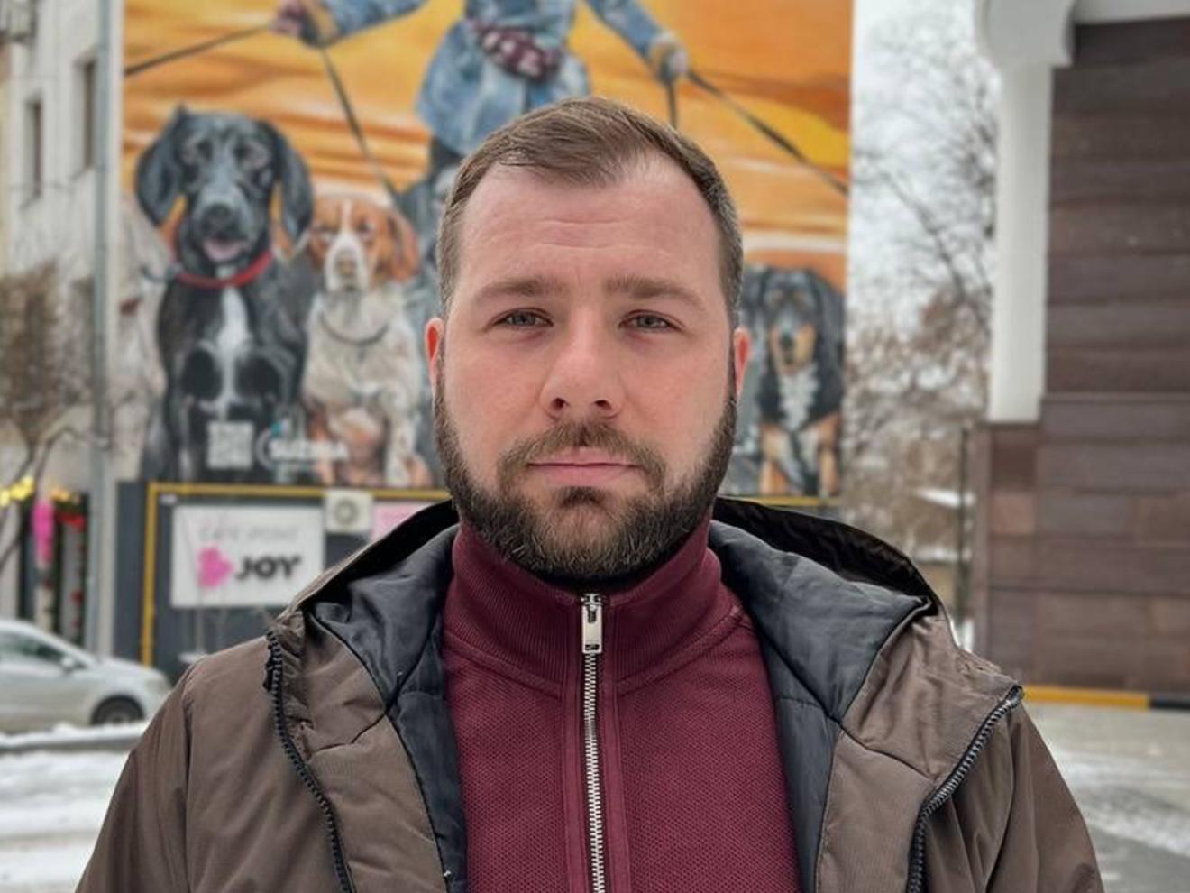 Ein Mann mit kurzem Bart und brauner Jacke steht vor einer bunten Wandmalerei, die eine Person mit mehreren Hunden an der Leine zeigt.