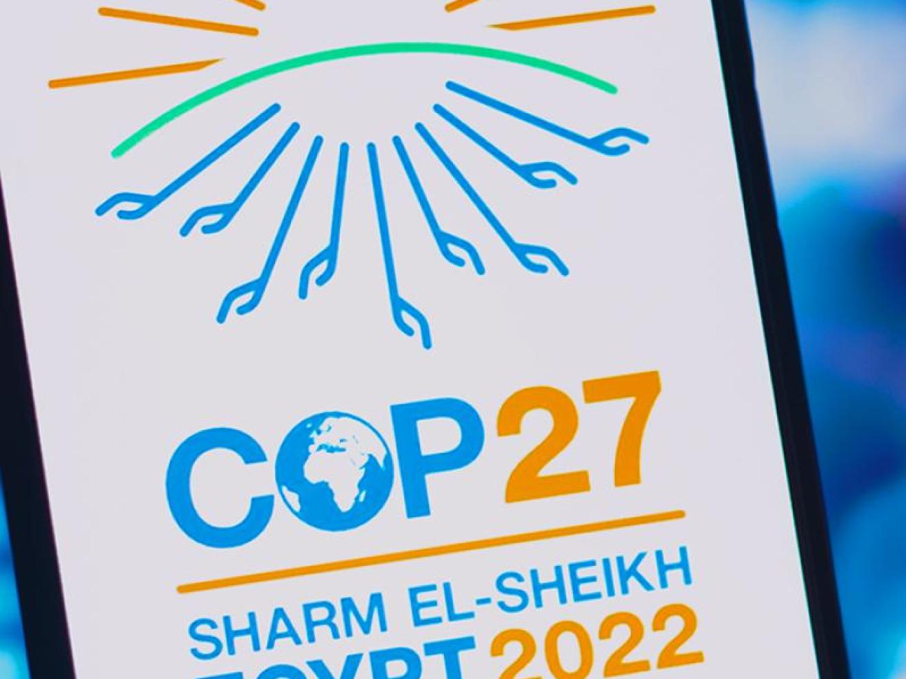 Ein Werbeplakat für die Klimakonferenz COP27 in Sharm El-Sheikh, Ägypten 2022. Das Design zeigt eine stilisierte Sonne in Orange und Grün über dem blauen Logo für COP27, darunter der Globus, und den Veranstaltungsort und das Jahr in Blau und Orange.