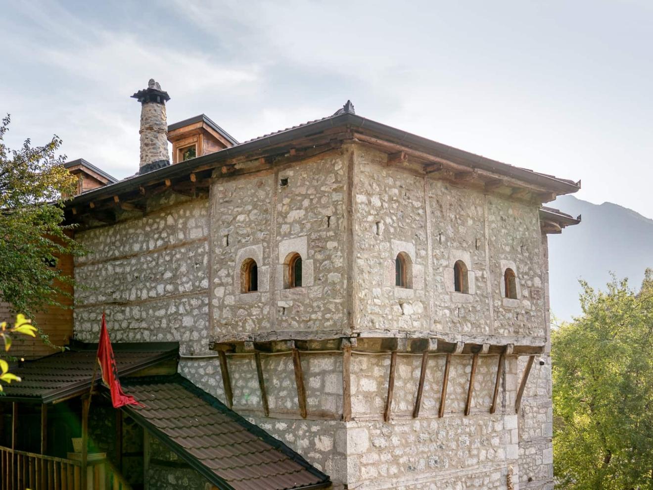 Ein traditionelles Steinhaus mit kleinen Fenstern, Holzdetails und der roten Fahne Albaniens steht inmitten grüner Bäume mit Bergen im Hintergrund.
