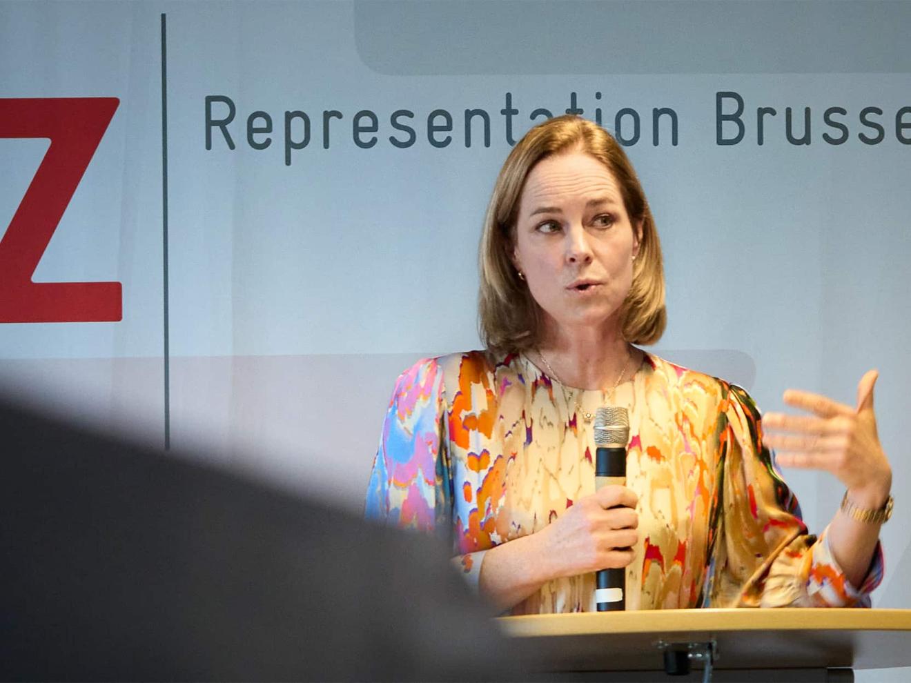 Eine Frau mit schulterlangem blondem Haar trägt eine farbenfrohe Bluse mit abstraktem Muster und spricht auf einer Veranstaltung in Brüssel an einem Rednerpult mit einem Mikrofon in der Hand.