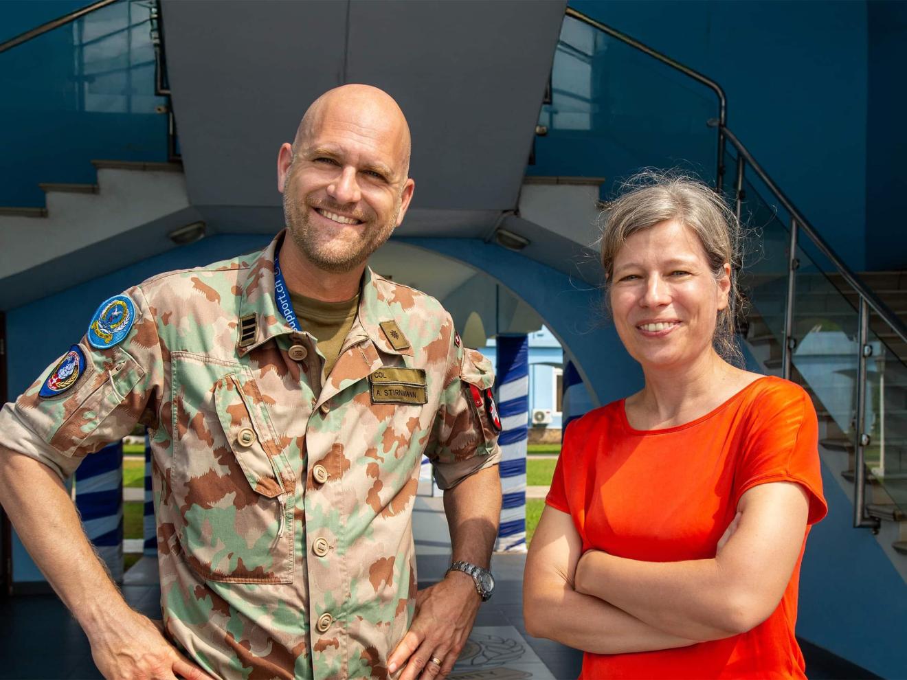 A smiling man in military camouflage uniform with a name tag reading ‘A. Stirnimann’ stands next to a woman in a red shirt in front of a modern blue building.