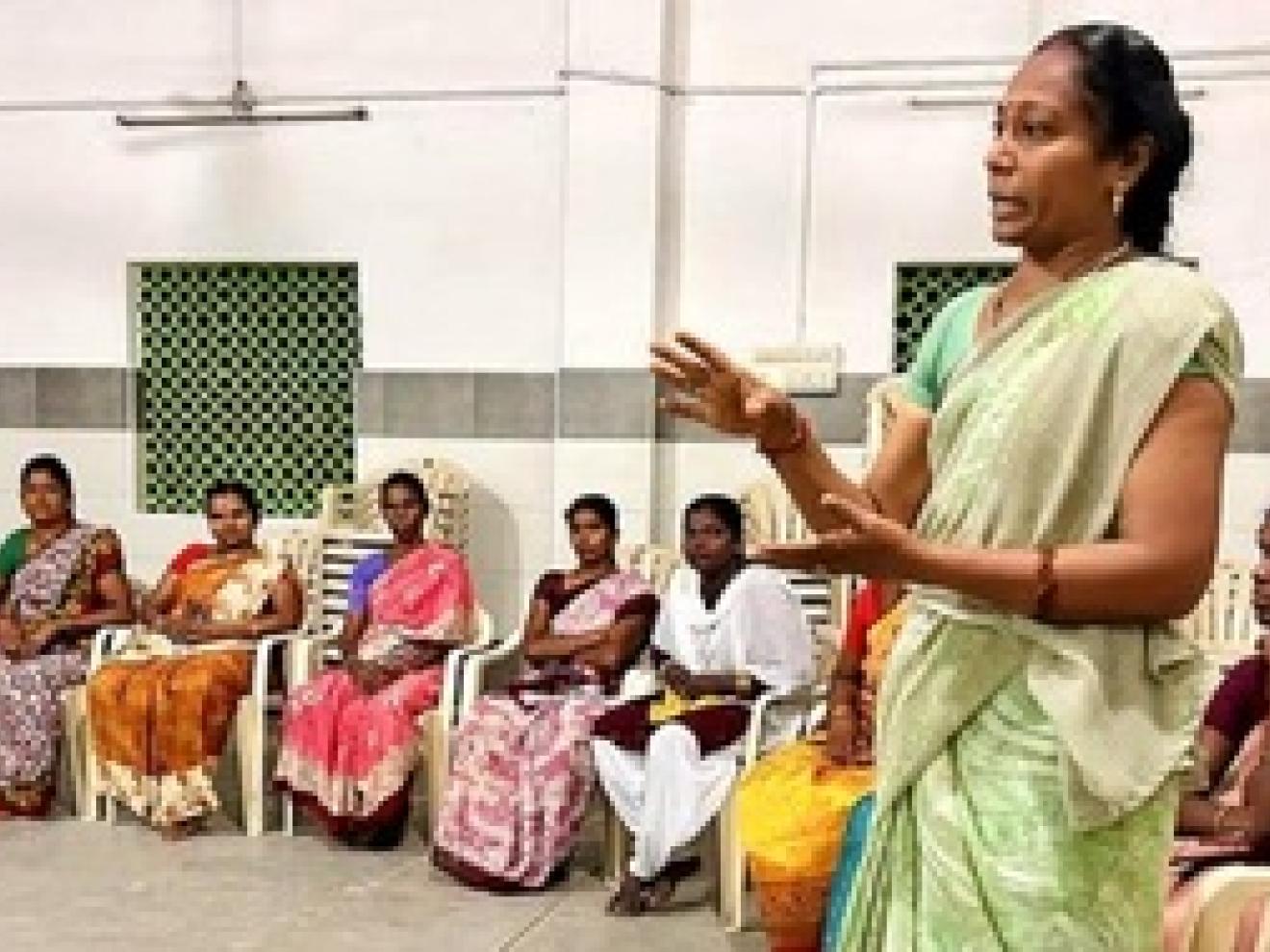 A woman stands in a circle of seated textile workers and explains something.Textile workers work in a factory.