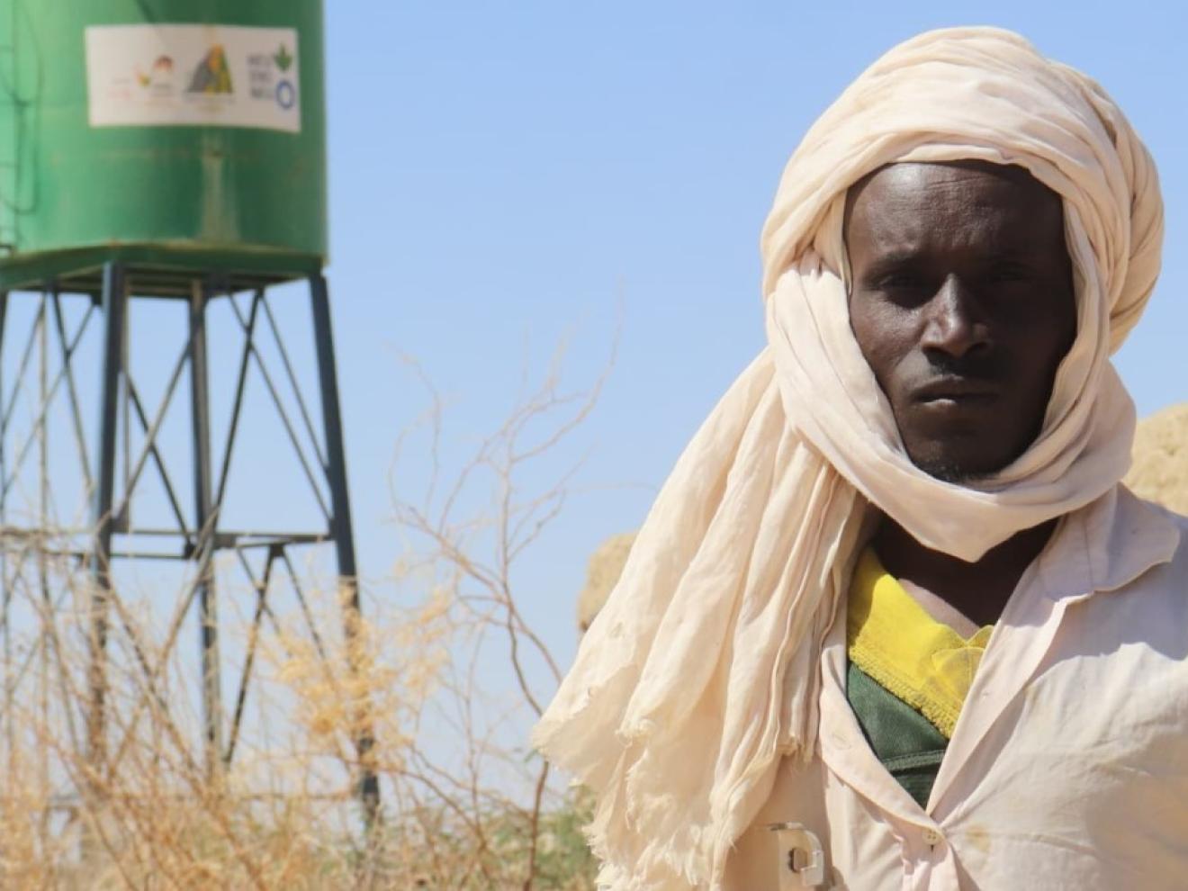 Ein Mann – Ibrahim Yatara – steht in Wüstenkleidung vor einem grünen Wassertank in einer trockenen Landschaft.