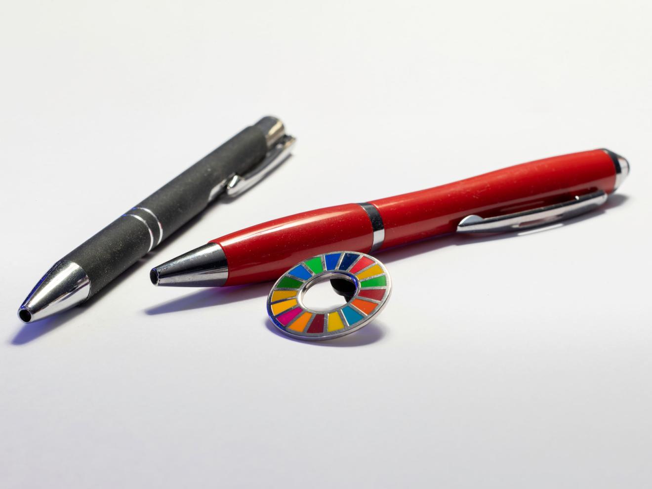 Zwei Kugelschreiber liegen auf einer weißen Oberfläche. Neben ihnen liegt ein kleiner runder Anstecker mit den Farben der SDGs. 
