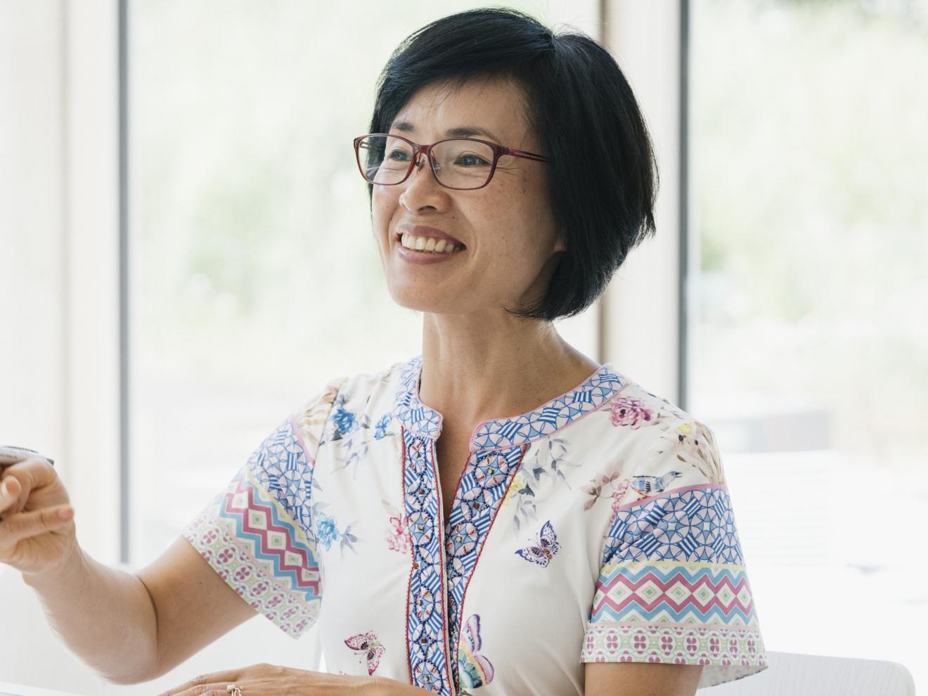 Eine lächelnde Frau gestikuliert mit einem Stift in der Hand. Sie sitzt in einem hellen Raum mit Blick ins Grüne.