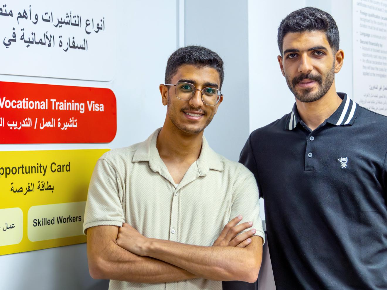Two young men stand side by side in front of an informational poster about visa and work opportunities in Jordan.