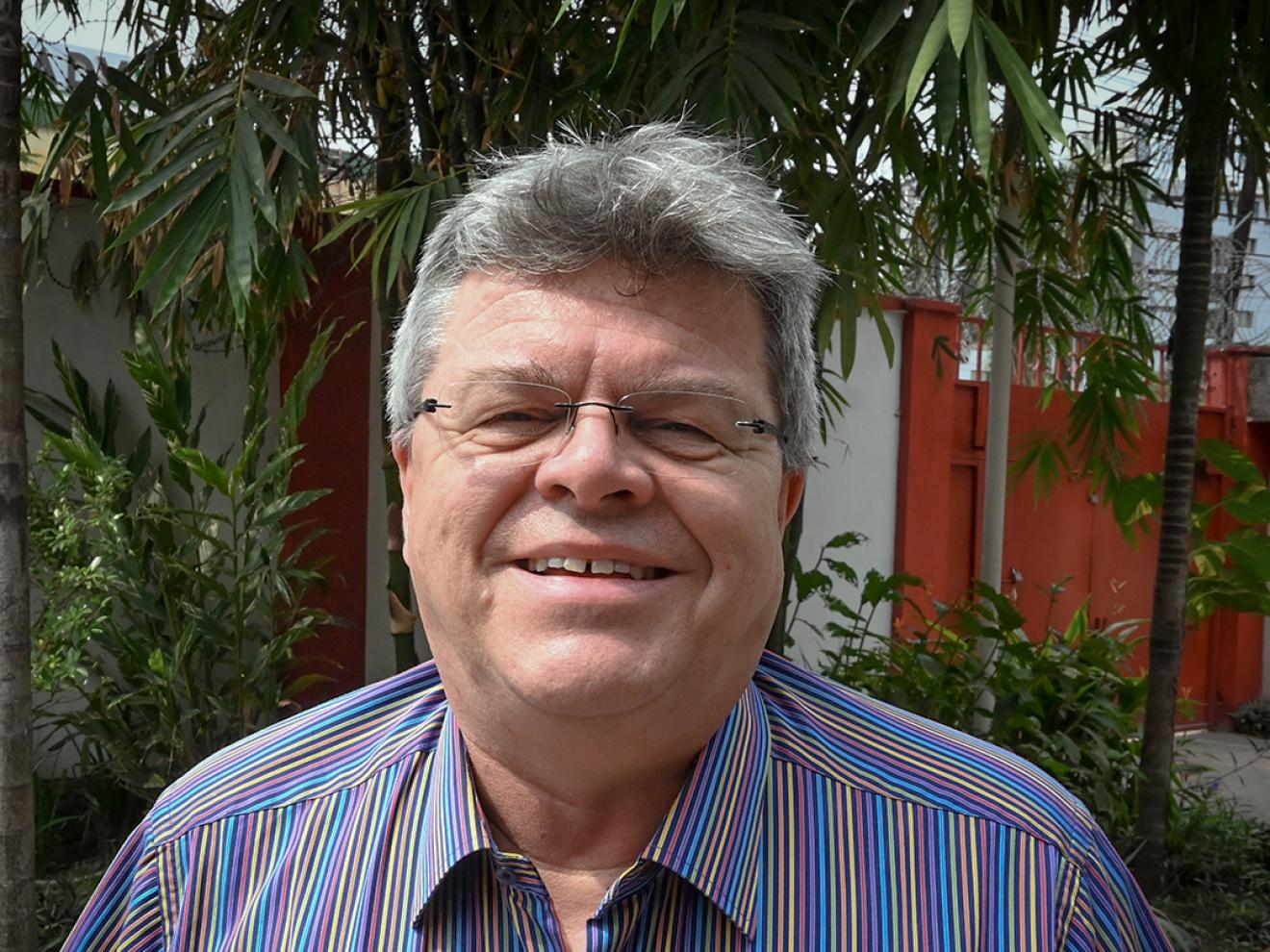 A man with gray hair and glasses smiles at the camera, wearing a brightly striped shirt and standing outdoors in front of tropical plants.