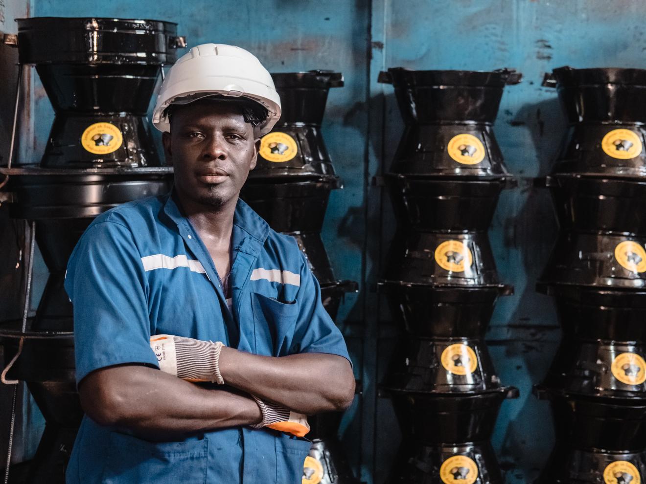 A man in a white helmet stands in front of a wall where cookers are piled up.