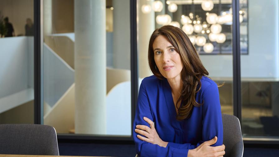 A woman with long brown hair wearing a blue blouse sits at a conference table in a modern office with glass walls and round ceiling lights in the background.