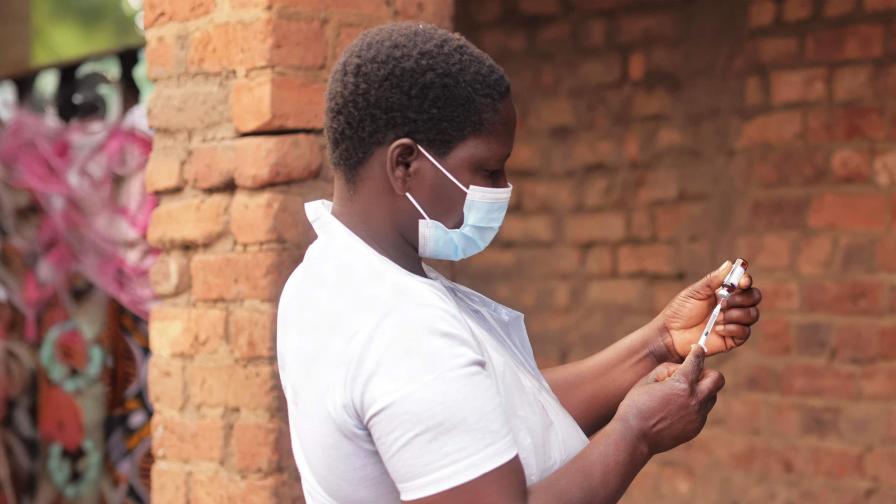 A woman wearing a medical face mask and a white T-shirt draws up an injection from a vaccine vial outdoors.