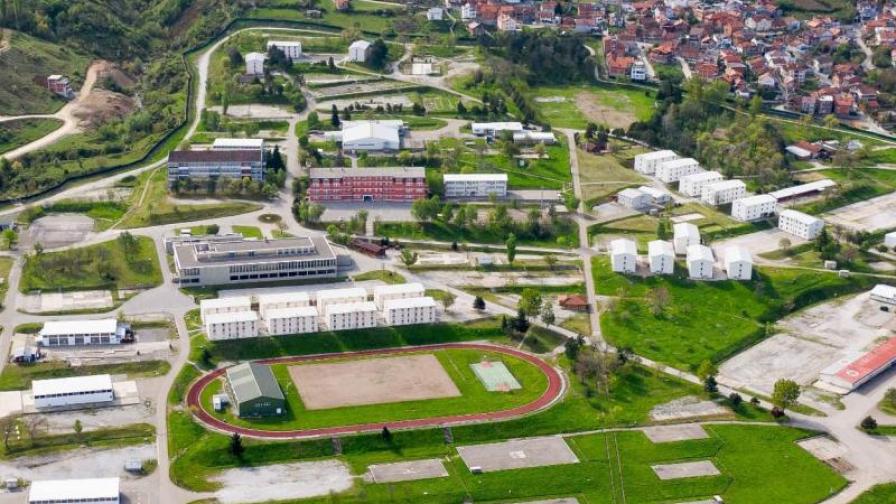 Luftaufnahme eines weitläufigen Geländes mit Gebäuden, Sportanlagen und einer angrenzenden Stadt in einer hügeligen Landschaft.