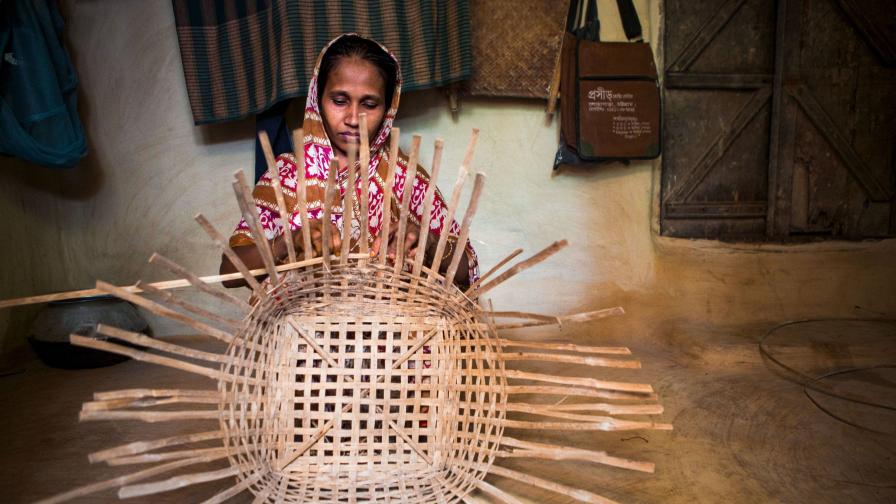 Eine Frau in traditioneller Kleidung sitzt in einem einfachen Innenraum und flechtet konzentriert einen grossen Korb aus Bambus oder ähnlichem Naturmaterial.       ChatGPT fragen