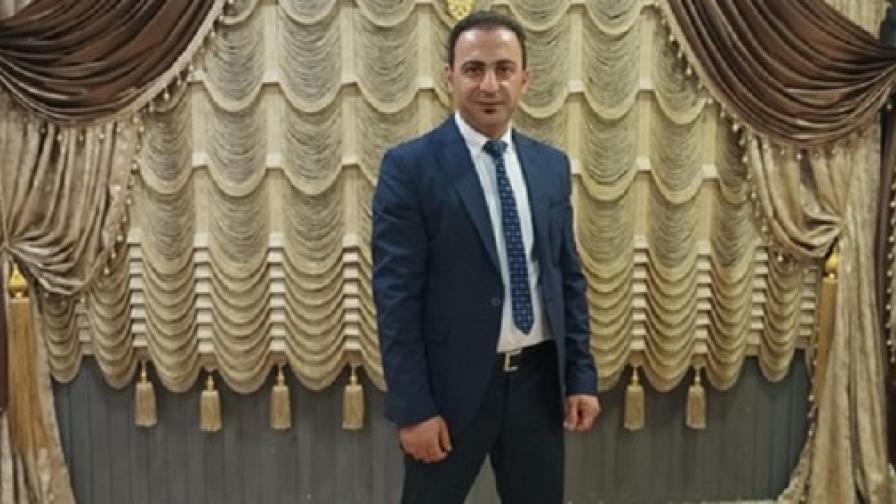 A man wearing a suit and tie is standing in front of artistically draped curtains in gold, beige and brown hues.