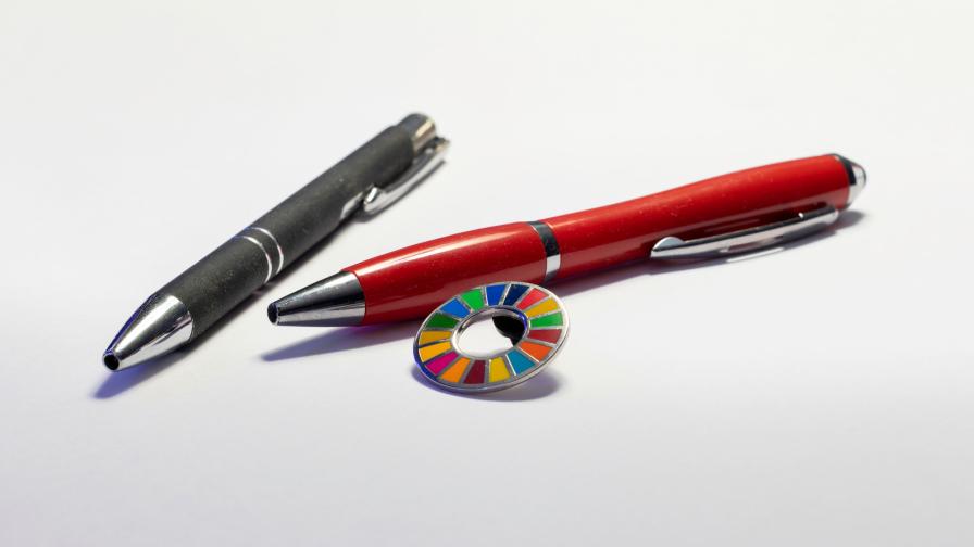 Zwei Kugelschreiber liegen auf einer weißen Oberfläche. Neben ihnen liegt ein kleiner runder Anstecker mit den Farben der SDGs. 