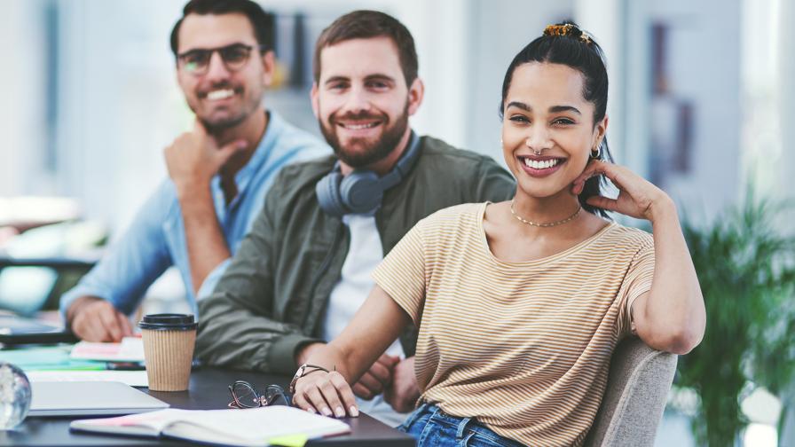 Drei junge Erwachsenen sitzen in einem Büro und schauen in die Kamera