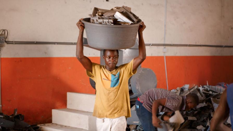 Zwei Männer vor einer weiß-roten Betonwand: Einer von ihnen beugt sich über einen Haufen aus ausrangierten Technikteilen, der andere trägt auf dem Kopf eine große Schüssel, gefüllt mit Technikteilen.