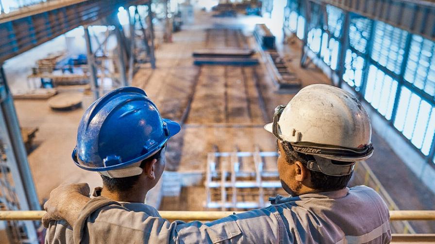 Two men in hard hats stand on a gallery looking down on a large industrial hall with machines and work areas.