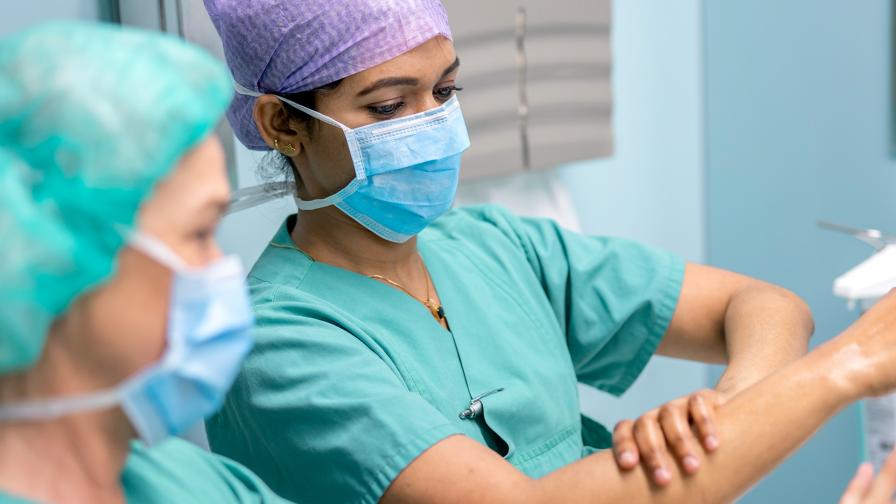 Two surgical staff members in green scrubs and face masks carefully disinfect their forearms.