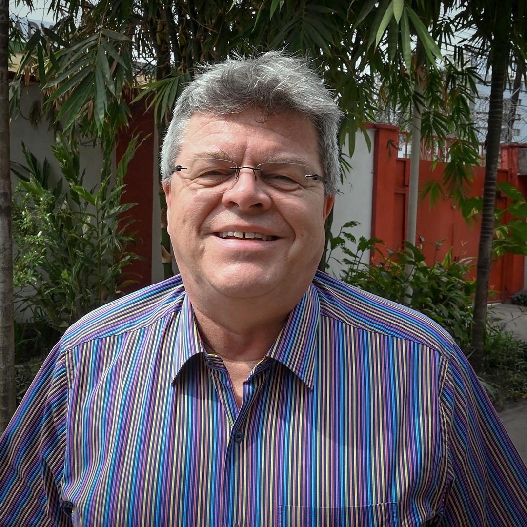 A man with gray hair and glasses smiles at the camera, wearing a brightly striped shirt and standing outdoors in front of tropical plants.