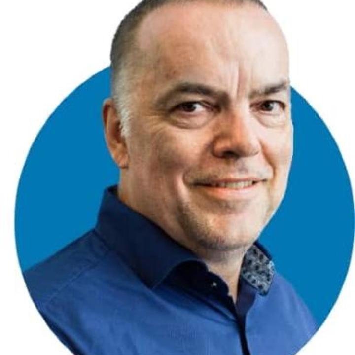 Porträt eines lächelnden Mannes mit kurzem Haar und blauem Hemd vor einem runden, blauen Hintergrund.