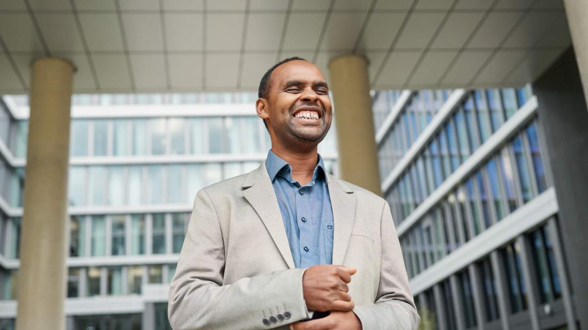 Ein lächelnder Mann in einem hellen Sakko und blauen Hemd steht vor einem modernen Bürogebäude mit Glasfassade.