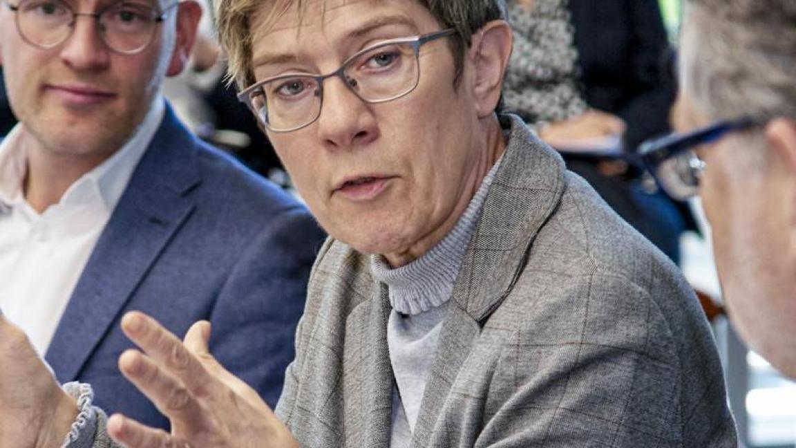 Diskussion in einem Konferenzraum mit einer Frau im grauen Blazer, die mit Gestik spricht, umgeben von mehreren Personen in Businesskleidung, die aufmerksam zuhören.