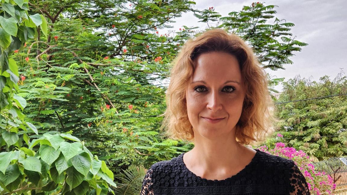 Eine Frau mit blonden, lockigen Haaren steht auf einem Balkon, trägt ein schwarzes, gemustertes Spitzenoberteil und lächelt leicht, im Hintergrund ist üppiges tropisches Grün mit Blüten und einem bewölkten Himmel zu sehen.