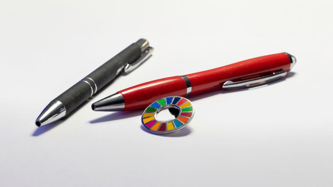 Zwei Kugelschreiber liegen auf einer weißen Oberfläche. Neben ihnen liegt ein kleiner runder Anstecker mit den Farben der SDGs. 