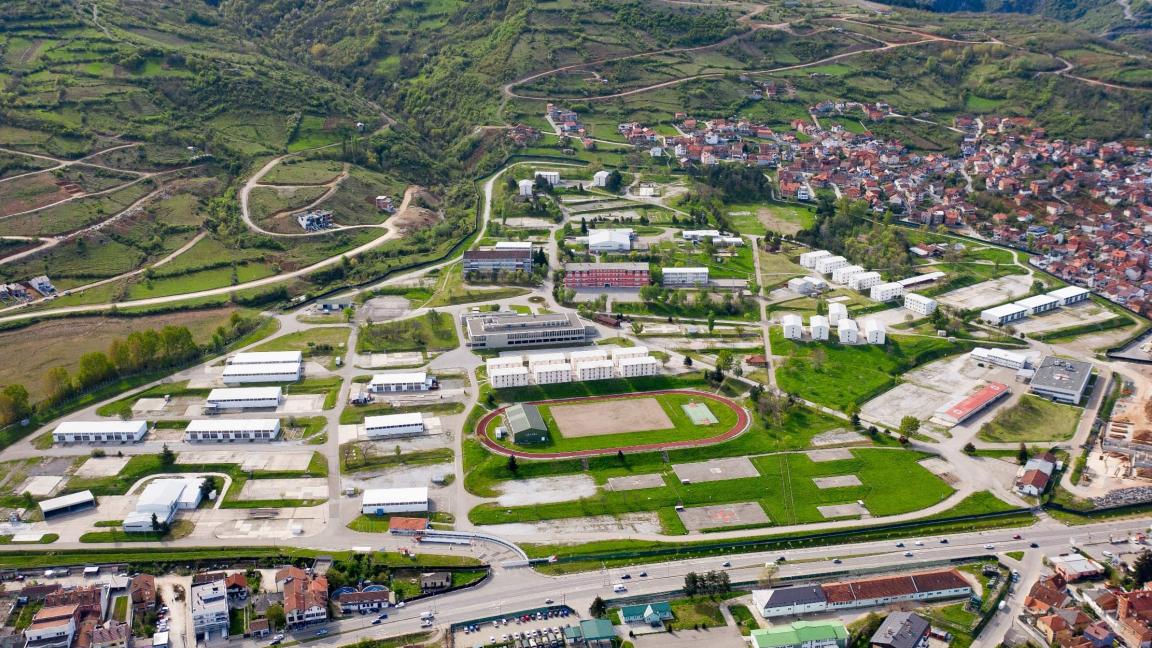 Luftaufnahme eines weitläufigen Geländes mit Gebäuden, Sportanlagen und einer angrenzenden Stadt in einer hügeligen Landschaft.