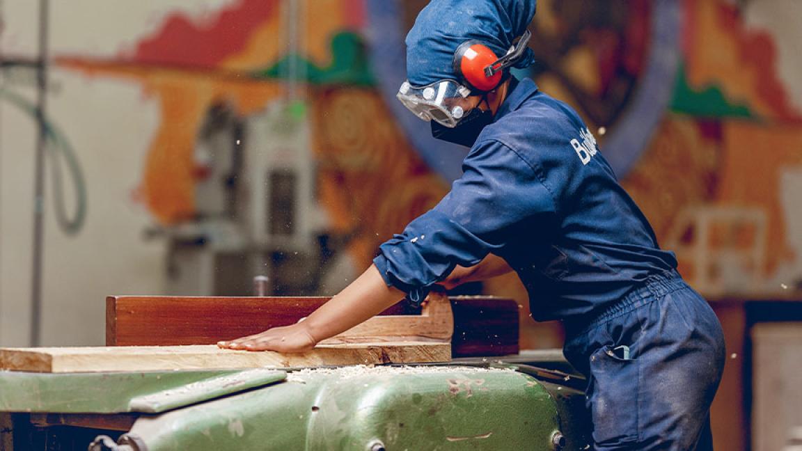 A woman in overalls and protective gear uses a woodworking machine in a workshop with a colourful wall mural.