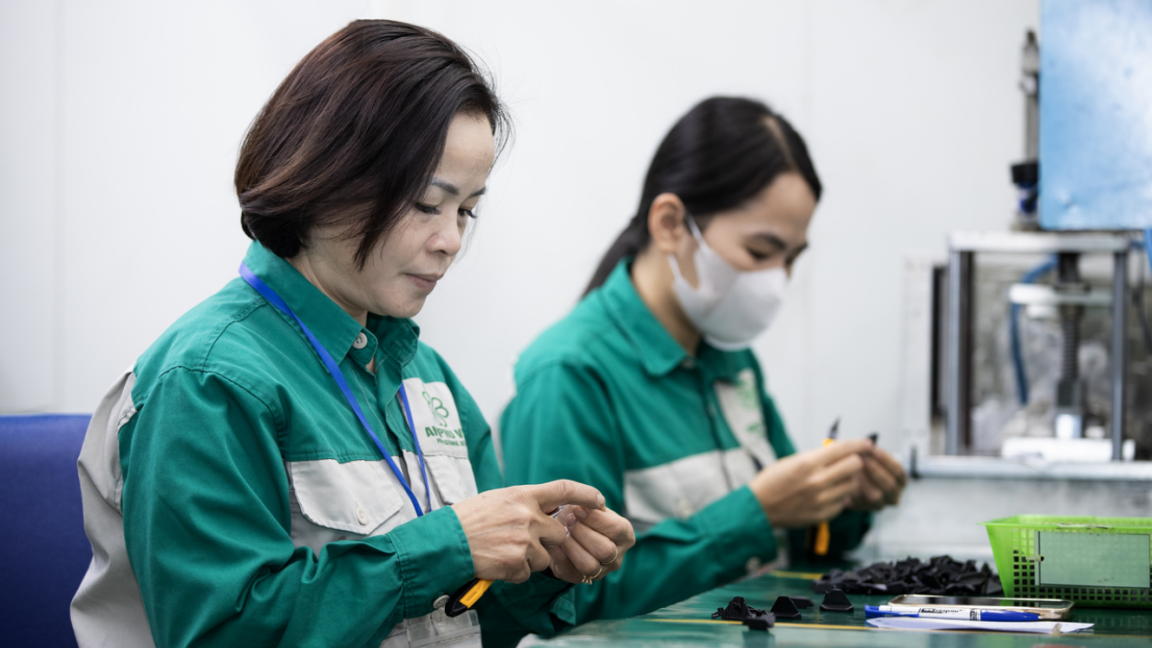 Zwei Frauen in Arbeitskleidung, die an einem Tisch sitzend kleine Plastikteile mit einem Schneidemesser bearbeiten. 